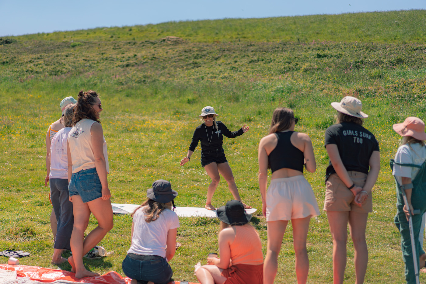 Learning to surf in the Summer sun, in one of Cornwall's best Surf Destinations. A group of women are on the grass being taught some theory by Allannah Brown