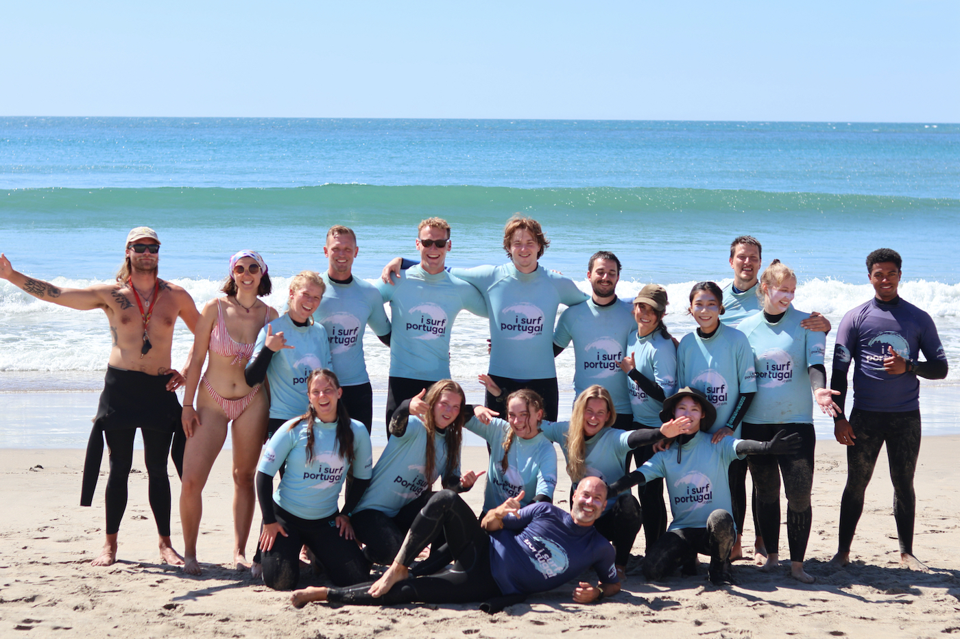 A group of students from I Surf Portugal on the beach infront of rolling Summer waves. Most of them are wearing wetsuits, there is one girl in a red swimsuit.