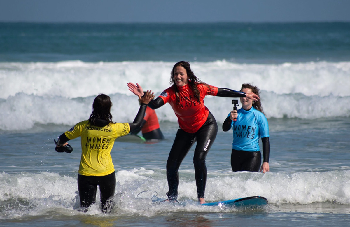 One women surfing high fiving another women, with another behind filming with a go-pro at a surf school in the UK